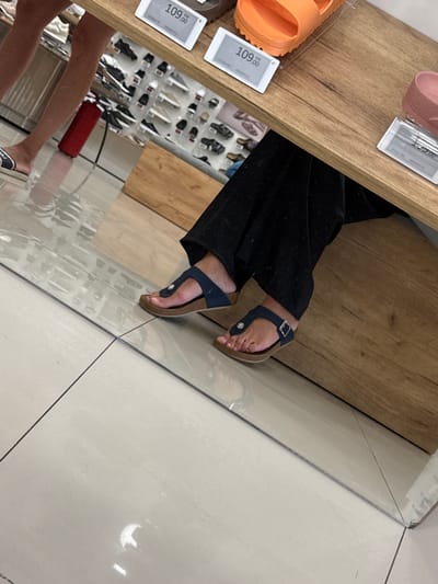 The image depicts a close-up view of a person's feet and legs, wearing dark pants and dark blue thong sandals, standing on a tiled floor. The person is positioned in what appears to be a retail environment, possibly a shoe store, as evidenced by display shelves filled with footwear visible in the background and price tags on a wooden surface in the foreground. One price tag displays "109,00 KM" and "BIRKENSTOCK". The floor is composed of large, light-colored tiles with dark grout lines. A reflection of the environment is visible on the floor. The scene is illuminated with artificial lighting, casting reflections and highlights. The location is identified as Ilidža, Bosnia and Herzegovina, suggesting this is a retail establishment within that city.