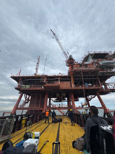 The image depicts a large offshore platform, likely situated near Marina di Ravenna, Italy, viewed from the deck of a supply vessel under an overcast sky. The immense, industrial structure, constructed from reddish-brown metal, dominates the left side of the frame. It features multiple levels, intricate piping, ladders, and various modules, along with two prominent industrial cranes extending into the cloudy sky.

The yellow, textured deck of the vessel occupies the right side, showing wear with black scuff marks. Several individuals, presumably workers, are present on the deck. In the foreground, a person is seen in a dark grey jacket, with a black bag or vest next to them clearly displaying the "HELLY HANSEN" logo (reversed) and "HH". Further along the deck, another worker wears a dark blue suit and a yellow hard hat, while a third is visible in an orange high-visibility vest. Additional figures can be seen in the distance on the deck.

Various personal belongings and equipment are scattered across the deck, including multiple travel bags, backpacks (one with a camouflage pattern), industrial cases, a yellow hose, and white boxes. A coil of blue rope rests on the deck. On a white sign affixed to the platform structure, the text "PCW-C" is discernible. The scene captures a moment during daytime, suggesting an operational transfer or arrival of personnel and cargo at the offshore facility, with the weather appearing cloudy and muted.