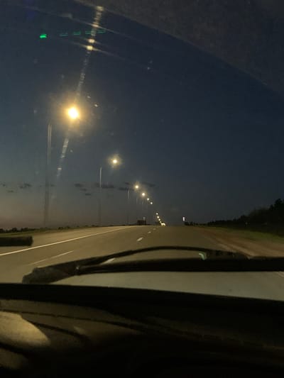 The image depicts a highway at dusk, viewed from inside a vehicle. The sky is a deep blue transitioning to a lighter hue near the horizon, with scattered clouds. A line of streetlights illuminates the road, casting a warm glow. The road itself is paved, with white lane markings visible. To the left of the road, there is a grassy embankment. To the right, a treeline is visible in the distance. The foreground shows the interior of the car, including the windshield wipers. The scene is devoid of any people or animals. The overall atmosphere is one of quiet travel on an open road at twilight.