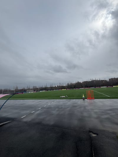 The image shows a view of a soccer field and surrounding area in Charentsavan, Armenia on a cloudy, overcast day. The field is green artificial turf, with white lines marking the boundaries. Orange cones and other training equipment are placed on the field. A stack of soccer balls is visible. Advertising boards line the side of the field. The foreground is a wet, paved surface, possibly a running track or walkway. Beyond the field, there are trees and buildings, and utility poles are visible in the distance. The sky is a uniform grey, indicating overcast weather.