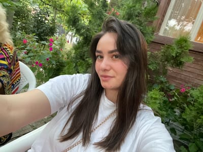 The image captures a young woman with long, dark brown hair and fair skin, looking towards the camera with a slight smile. She is wearing a white t-shirt with a gold chain strap visible across her chest. The background features lush green foliage, including trees and pink flowers, indicative of an outdoor setting. A wooden building with a window is partially visible behind the greenery. The overall scene suggests a casual outdoor gathering or a moment of relaxation in a garden environment. The location is identified as Simitli, Bulgaria.
