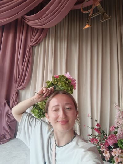 A young woman with blonde hair and blue eyes is taking a selfie, winking at the camera. She is wearing a light grey t-shirt and has on hoop earrings. She is holding a bouquet of pink and green artificial flowers above her head. The background consists of draped pink and beige curtains, suggesting an indoor setting possibly decorated for an event. A decorative, golden letter "A" is visible hanging from the ceiling in the top right corner. The lighting suggests it might be daytime. The woman appears happy and playful. The location is identified as Starobzhegokay, Russia.