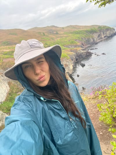 A medium-shot selfie shows a young woman with dark hair wearing a grey bucket hat and a blue hooded waterproof jacket. She is winking and making a kissing face. The background reveals a rugged coastal landscape. To the left, a grassy slope leads down to a rocky coastline and the ocean. Vegetation is also visible on the right side of the frame. The scene is in Gvozdevo, Russia.
