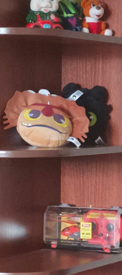 This is a vertical, medium-shot image of three shelves in a wooden bookcase. The bookcase is filled with various toys.
On the top shelf, from left to right, there is a ceramic pig figurine riding a red motorcycle, a green bag, and a brown teddy bear wearing a red shirt.
The middle shelf contains two stuffed toys. The one on the left is orange and appears to be a character with large purple and yellow eyes and an open mouth showing jagged teeth. The toy on the right is black and also has a green eye. Both toys have white tags attached to them.
The bottom shelf holds a red and yellow remote-controlled helicopter inside a clear plastic case. The case has red latches on the bottom. A yellow label with black text is visible on the helicopter.
The overall environment appears to be an indoor setting, possibly a child's room or a display area. The lighting is dim, and the image quality is slightly grainy. No specific geographical markers indicating Aktanyshbash, Russia, are present in the image itself.
