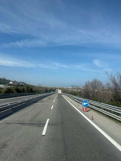 The scene depicts a multi-lane highway, likely within the vicinity of Martinsicuro, Italy, under a clear blue sky with sparse, wispy cirrus clouds. The perspective is forward-facing from the rightmost lane of travel. The asphalt road surface features white dashed lane markings in the center and a solid white line defining the right shoulder. A metal guardrail borders the right side of the highway, behind which an embankment with dry, deciduous trees and low-lying vegetation is visible. Positioned on the right shoulder, approximately mid-frame, is a blue circular road sign displaying a white left-pointing arrow, mounted on an orange A-frame stand. Several small orange traffic cones are arrayed near the sign and along the shoulder. In the distance, a white recreational vehicle or large white truck is visible ahead in the same lane, followed by other indistinct vehicles. To the left of the main travel direction, multiple highway lanes are separated by a concrete median barrier. A verdant hillside with scattered, indistinct buildings rises on the far left. The distant horizon reveals an urban or semi-urban landscape, consistent with a coastal setting, under a light atmospheric haze. No individuals are visible.