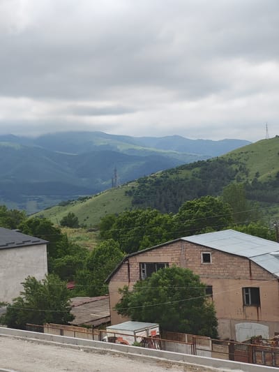The image depicts a rural mountainous landscape in Bazum, Armenia, under a cloudy sky. The foreground shows a dirt road or path with a concrete barrier and a chain-link fence. Behind the fence, several buildings are visible. To the left is a light-colored building with a dark, sloped roof. In the center and right, a tan-colored brick building with a corrugated metal roof is prominent. This building has several windows, some of which appear broken, and exposed brickwork. Smaller, older-looking structures with grey, weathered roofs are also present behind the main buildings.

Beyond the buildings, a dense forest of green trees stretches across the middle ground. The background is dominated by rolling, lush green mountains that recede into the distance. The peaks of the farthest mountains are partially obscured by mist or low-lying clouds. A tall, metallic transmission tower stands on a green hillside in the mid-background, indicating the presence of infrastructure in the remote area. The overall atmosphere is somewhat somber due to the overcast weather, suggesting it is likely daytime but not a bright, sunny period. There is no visible text in the image.