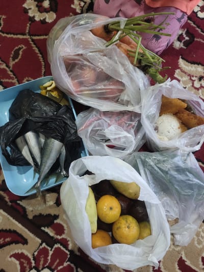 In Cintadamai, Indonesia, on a patterned rug, a selection of groceries are laid out. A light blue tray contains several whole fish, wrapped in black plastic, with slices of citrus fruit nearby. Several plastic bags hold a variety of produce, including carrots with green tops, oranges, avocados, and what appears to be some type of green vegetable. Another bag contains rice and what seem to be fried items. A person's lap, clad in pink patterned fabric, is partially visible in the upper right corner, suggesting they are the owner of the groceries. The scene appears to be indoors, possibly a home, with the focus entirely on the assortment of food items.