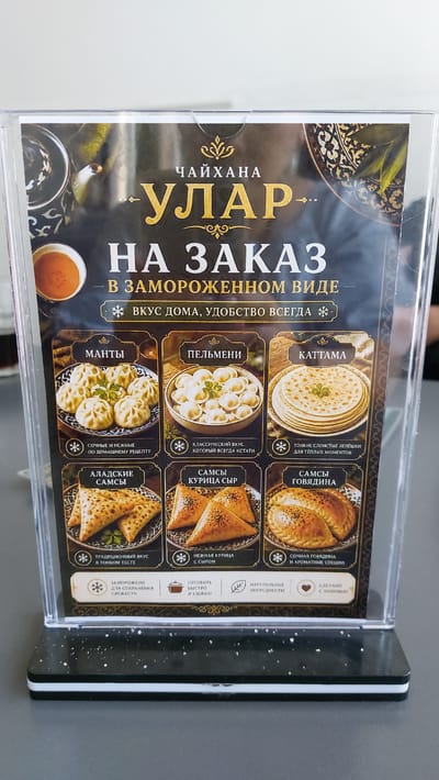 The image shows a menu board for a restaurant called "ЧАЙХАНА УЛАР" (Chaikhаna Ular). The menu is in Russian and features frozen food items available for order. The board is divided into sections, each showcasing a different dish with a picture and a brief description.
The dishes featured are:
* **KATTAMA:** Thin layered flatbreads for warm moments.
* **PELMENI:** Classic taste, always appropriate.
* **SAMSY GOVYADINA:** Juicy beef with aromatic spices.
* **MANTI:** Juicy and tender, according to a homemade recipe.
* **SAMSY KURITSA SYR:** Delicate chicken with cheese.
* **ALADSKIE SAMSY:** Traditional taste in thin dough.
Below the main menu items, there are also general descriptions: "Frozen to preserve freshness," "Natural ingredients," and "Made with love," as well as "Convenient ordering, comfort at home." The overall theme of the menu is convenient, homemade-style food that can be enjoyed at home, even when frozen.
The menu board itself is presented in a clear plastic holder, suggesting it might be a freestanding display or a tabletop sign. The background of the menu features dark, ornate patterns reminiscent of Central Asian textiles, with images of food items like dumplings, flatbreads, and savory pastries. The lighting suggests it's indoors, likely in the restaurant itself. The overall presentation is professional and aims to highlight the quality and convenience of the restaurant's offerings.