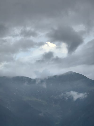 The image shows a wide shot of misty mountains. The sky is overcast with dark gray clouds, with a small patch of bright light breaking through in the upper center. A large, dark mountain range dominates the lower half of the frame. Clouds and mist are clinging to the slopes, obscuring some of the details of the terrain, which appears to be covered in dark green vegetation and patches of lighter green. A small bird or insect is visible in the sky above the mountain. The overall atmosphere is moody and dramatic, suggesting a cool, possibly damp climate. There are no people or man-made structures visible in the image. The location is described as Siumsa, India.