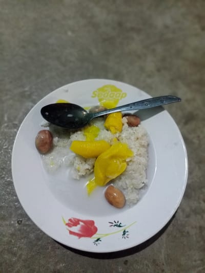 A plate of sticky rice with jackfruit and peanuts is shown, with a spoon resting on it. The plate is white with a floral design and the word "Sedaap" printed on it. The food appears to be in a dish of coconut milk. The plate is placed on a textured surface, possibly a table or countertop. The lighting suggests the image was taken indoors or at night.