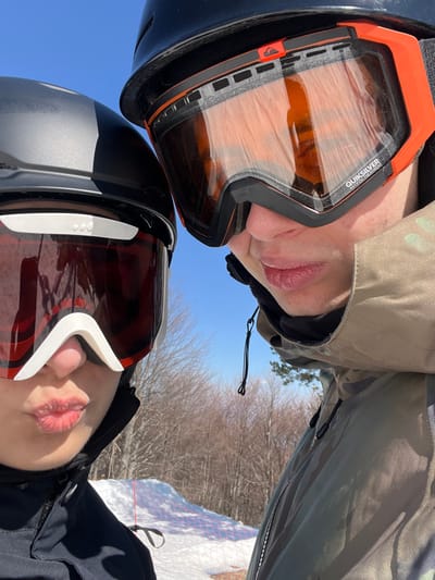 A close-up, low-angle image captures two individuals on a ski slope in Bogoslov, Bulgaria, under a clear blue sky. The person on the left wears a matte black ski helmet and dark red-tinted ski goggles with a prominent white nose bridge, their lips visibly puckered in a "kiss face." A portion of a black winter jacket is visible. The person on the right wears a black ski helmet with an orange top accent and orange-tinted Quiksilver ski goggles, their lips displaying a neutral expression. They are dressed in a camouflage-patterned winter jacket. The background reveals a snow-covered slope, a red safety net, and a line of bare deciduous trees under bright sunlight.