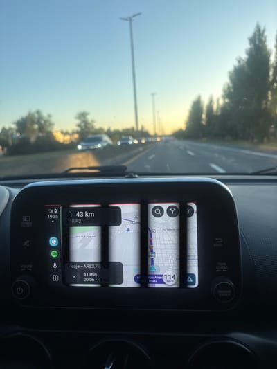 Inside a car, a digital dashboard display is visible. The top screen shows a map with a speed of 114 km/h towards Buenos Aires, Argentina. Below, the navigation indicates 43 km remaining and an estimated 31 minutes to destination. The ETA is 20:06. The route involves a toll costing approximately ARS 3,722.00. The time is 19:35. The car appears to be driving on a multi-lane highway with other vehicles present. The weather seems bright with a clear sky and trees lining the sides.