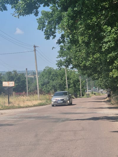 A silver sedan is driving down a paved road in Pidhaitsi, Ukraine, on a clear day. The road is lined with trees and utility poles. A person is jogging on the right side of the road. In the background, there are fields and more trees. The car's license plate is partially visible and reads "BA 3350 BX".