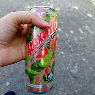 A hand holds a can of "Mojito" flavored non-alcoholic beverage. The can features a vibrant design with images of strawberries, limes, and mint leaves, suggesting a refreshing summer drink. The text on the can is in Russian, indicating the brand "Ochakovo" and the flavor "Mojito Strawberry" as a non-alcoholic, medium-carbonated refreshing cocktail. A "Halal" certification logo is also visible. The background appears to be a textured, grayish surface, possibly asphalt or gravel, with dappled sunlight indicating an outdoor setting during the daytime, likely a warm day. The image focuses on the product, highlighting its visual appeal and key characteristics.