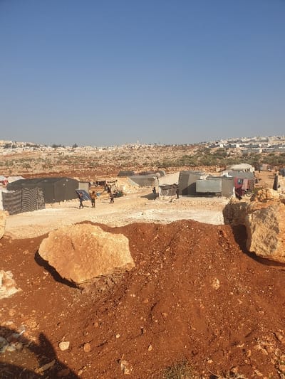 The image, when correctly oriented, presents a wide view of an arid landscape in Killi, Syria, under a clear, bright blue sky. The foreground is dominated by reddish-brown, rocky earth and large, light-colored boulders, indicating a rugged and uneven terrain. A dirt path or cleared area runs through the midground, alongside a series of makeshift shelters or tents. These structures vary in appearance, with some featuring blue tarpaulins, suggesting an informal settlement or camp.
Several indistinct human figures are visible near these shelters, but their specific activities are not clearly discernible, appearing to be engaged in daily camp life. In the far background, a sprawling town or village can be seen on a distant hillside, with numerous buildings extending across the landscape, interspersed with sparse vegetation. The strong, defined shadows cast by the rocks and shelters indicate bright daylight, suggesting it is either mid-morning or early afternoon. The weather is clear and sunny with no clouds visible. There is no discernible text within the image. The overall scene conveys the reality of an informal settlement in a harsh, rocky environment.