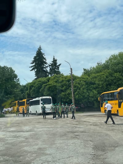 The scene is an outdoor area, likely a parking lot or a gathering space, in Tsar Kaloyan, Bulgaria. The sky is partly cloudy. The ground is concrete. A line of people in dark green uniforms and light green caps stands in formation. Some are holding green flags or poles. In the background are buses, some are yellow, some are white. One man is standing by a yellow bus wearing a white shirt and dark pants. A line of trees runs across the center of the image. A utility pole stands among the trees.