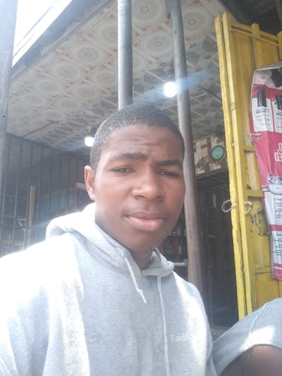 A young, dark-skinned male with short, dark hair, wearing a light grey hooded sweatshirt, is captured in a head-and-torso portrait. He faces the camera directly with a neutral expression. The scene is stated to be in Igwuruta, Nigeria.
In the immediate foreground, three prominent dark, vertical metal bars partially obstruct the view, one positioned near the subject's left shoulder, another near his right, and a third further to the left.
The background features a light-colored, patterned ceiling with repeating circular designs in red and white. Two bright, exposed light bulbs hang from the ceiling, producing significant lens flare.
To the left of the subject, behind the vertical bars, a dark, possibly corrugated metal structure is visible. Beyond this, an interior space with shelving containing various items is dimly visible.
To the right of the subject, a weathered yellow metal frame or door jamb with signs of rust is present. Attached to this frame is a white and red commercial banner or poster with partially legible text. Behind the yellow frame and banner, another interior space with shelves and a partially visible green logo (resembling "OPPO") is discernible. The overall lighting is bright, combining natural and artificial light sources.