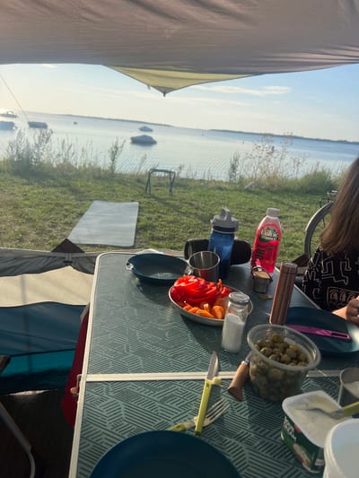 In Am Salzhaff, Germany, a table is set for a meal at a campsite. A teal and gray patterned table holds a bowl of sliced red bell peppers and carrots, a salt shaker, a clear plastic container filled with green olives, and a tub of sour cream. A metal water bottle, a pink "Volvic" drink bottle, and a metal mug are also on the table. To the right, a person's shoulder and arm, wearing a black shirt with a white geometric pattern, are visible. Behind the table, a bicycle is parked. The campsite overlooks a body of water with several boats moored, and a grassy area with a mat is visible in the background. The scene is illuminated by natural daylight, suggesting it is either morning or late afternoon.