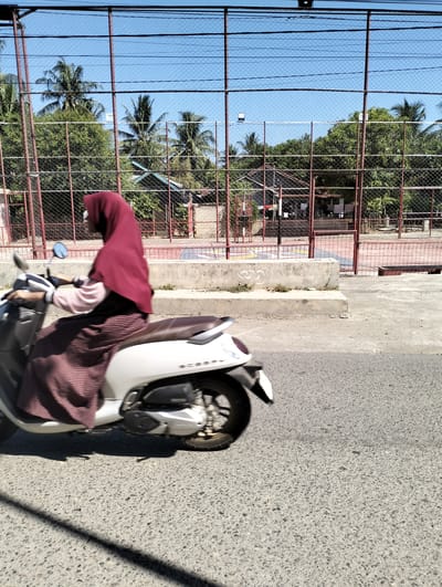A woman wearing a burgundy hijab and a patterned skirt rides a white scooter on a paved road. The scooter has a brown seat and the word "SCOOPY" is visible on its side. In the background, a fenced-in sports court with a red surface and white markings is visible. Trees, including palm trees, and a few buildings are also present behind the court, under a clear blue sky. The image appears to be taken during the daytime. The specific location mentioned, "Janggotseungko, Indonesia," is not explicitly identifiable from the visual elements in the image.