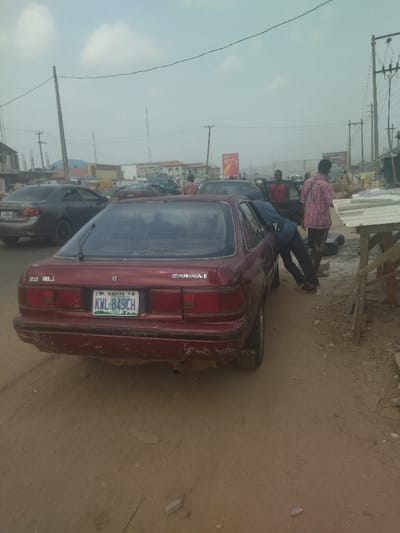 The image shows a red Toyota Carina II sedan parked in a dusty street in the city of Warri, Nigeria. The car is parked on the right side of the road, facing the camera. There are several other cars parked on the left side of the road, as well as a few people walking in the background. The car is surrounded by a small amount of debris and loose sand. The license plate reads KWL-849CH. The road is unpaved, dry, and dusty. There are a few power lines running over the road and across the background. There are several buildings visible in the background, with advertising signs on them. There are two men interacting, with one man wearing a blue shirt and jeans leaning down and looking at the car and another man standing upright next to the car, wearing a red and white patterned shirt. The scene is taking place in a residential or commercial district of Warri, Nigeria.
