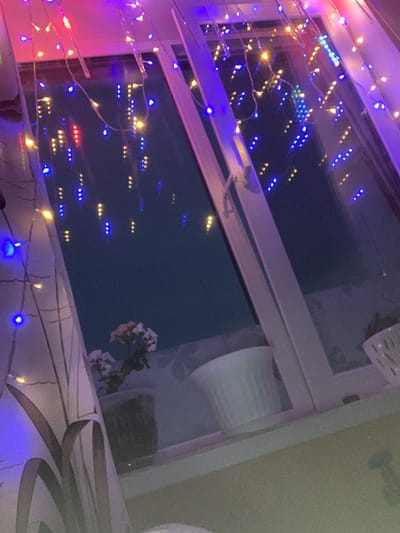 This media file captures a close-up view of a window decorated with colorful LED string lights, creating a festive and cozy atmosphere. The lights, predominantly blue and yellow, with some red and pink hues, are draped along the window frame and appear to be reflected in the glass. The scene suggests an indoor setting, likely a home, with the window looking out onto a dark exterior, implying it is nighttime or dusk.
In the reflection on the window, a faint pattern of lights can be seen, possibly from other buildings or streetlights in Leninogorsk, Russia. A few sparse, white flowers on a branch are visible in the upper right portion of the reflection, adding a delicate natural element to the otherwise artificial display of lights. The lighting is warm and inviting, with the bright LED lights casting colorful glows on the surrounding surfaces. The overall mood conveyed is one of cheerfulness and tranquility, characteristic of holiday decorations or a personal touch to an living space. No visible text is present in the image.