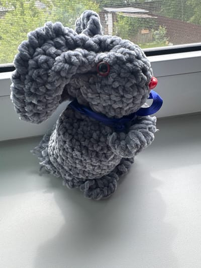 A handmade, gray, crocheted bunny sits on a white windowsill, facing right. The bunny has large, floppy ears, round red eyes, and a red nose. A blue ribbon is tied around its neck. Behind the bunny, a window with a screen is visible, showing greenery and part of a building outside. The overall setting appears to be indoors, near a window. The bunny is the sole subject of the image.