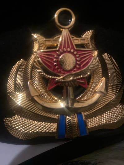 The image shows a close-up of a gold-colored emblem, likely a military or naval insignia, set against a black background. The emblem features a prominent red star with radiating lines and a circular emblem in its center. Below the star, a golden anchor is visible, intertwined with what appears to be a wreath or stylized laurel leaves. The bottom of the emblem has two blue rectangular elements. The lighting creates strong highlights and reflections on the metallic surface of the emblem. The location in Жданов ауылдық округі, Kazakhstan is not depicted in this image, as the scene is an isolated emblem.