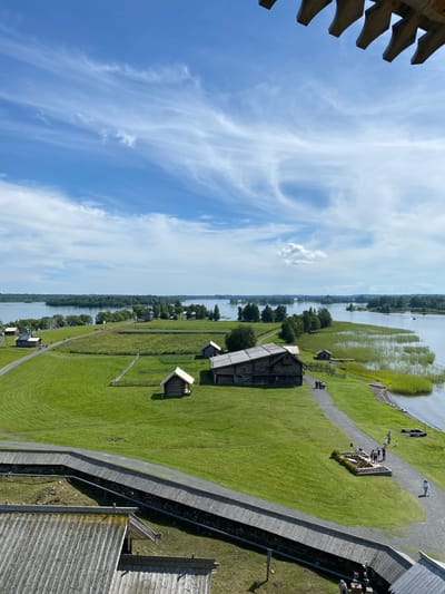 An aerial, eye-level, wide shot captures a historic village with wooden structures and a vast body of water under a bright, partly cloudy sky. The scene is located in Boyarshchina, Russia. In the foreground, a wooden walkway with a railing is visible, leading the eye into the expansive green landscape. Scattered across the grassy fields are several traditional wooden buildings, some appearing to be houses or barns, with thatched or wooden roofs. A larger, prominent wooden structure with a dark roof and visible windows stands towards the center-left of the frame. A gravel path winds through the village, and several small groups of people are seen walking along it, appearing to be tourists. The path leads towards the water's edge, where reeds grow thick along the shore. The calm water stretches out to a horizon dotted with distant, tree-covered islands. The sky above is a vibrant blue, with wispy white clouds creating texture and depth. The overall impression is of a serene, preserved historical site surrounded by natural beauty.