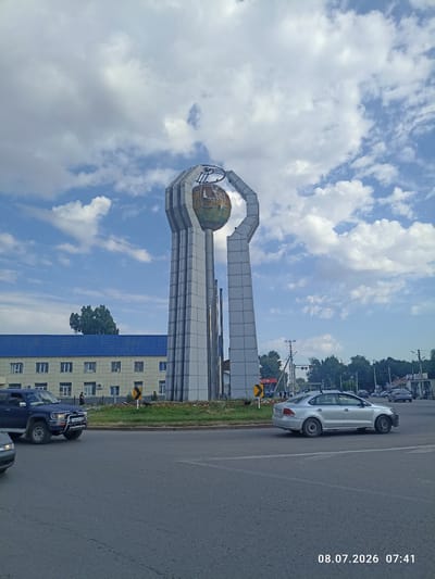 A wide shot captures the monument of Uzynagash, Kazakhstan, featuring a towering structure with two large, curved arms supporting a spherical globe. The monument is set against a backdrop of a bright blue sky filled with scattered white clouds. To the left of the monument, a long, light-colored building with a blue roof stands. A dark blue SUV is parked on the left side of the road in front of this building. Several cars are visible on the asphalt road in the foreground and background, suggesting an urban environment. Trees and smaller buildings line the horizon behind the monument, with power lines crisscrossing the sky. A timestamp in the bottom right corner reads "08.07.2026 07:41". The scene appears to be a daytime shot on a clear day.