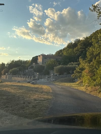 A paved road curves through a grassy, sun-drenched landscape towards a stone building situated on a hillside amidst lush greenery. The building, with a reddish-tiled roof and multiple windows, is partially obscured by trees. A stone wall with a gated entrance borders the property, and several large rocks are scattered on the grass. The sky above is a clear blue with scattered white clouds, illuminated by warm, late afternoon sunlight. The scene appears to be in Đenjaši Česminovo, Montenegro, based on the provided location context.