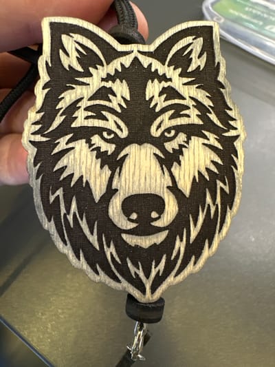 The image shows a close-up of a wooden pendant shaped like a wolf's head. The wolf's head is carved with intricate detail, with sharp lines and a textured appearance. The wood is light in color, and the carved areas are dark, creating a stark contrast. A black string is attached to the top of the pendant, suggesting it is meant to be worn as a necklace or keychain. A small, black cylindrical object is attached to the bottom of the pendant, from which a metal ring and clasp are visible. The background is out of focus, but appears to be a flat, dark surface. There is a blurred object in the upper right corner that appears to be a plastic case with some green text. The lighting suggests an indoor setting with a light source coming from the top. There are no indications of the city of Mikhailovka, Russia.
