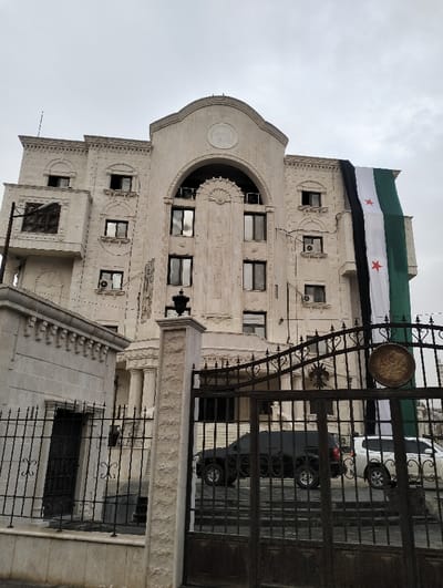 A light stone building with ornate details and arched windows is partially draped with a large Syrian flag. Wrought iron gates stand in front, partially obscuring two parked SUVs, one black and one silver. A smaller stone structure sits to the left, also behind a wrought iron fence. The sky is overcast. The building's architecture suggests an embassy or official government building.
