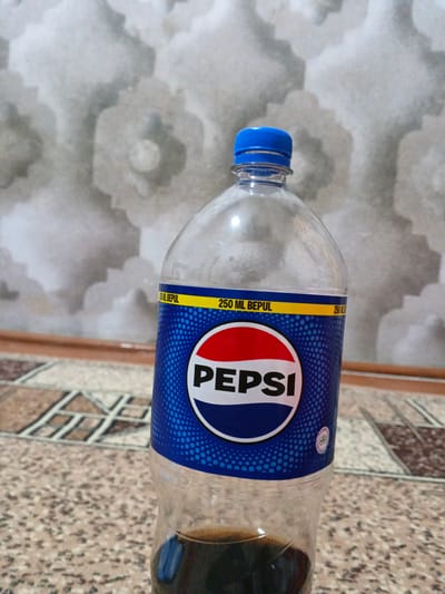 A medium shot shows a partially full plastic bottle of Pepsi, with a blue cap, standing on a patterned rug. The Pepsi logo, featuring the iconic red, white, and blue circle, is clearly visible on the blue label. The bottle is positioned in the foreground, with a blurred, textured wall serving as the background. The lighting appears to be natural, casting soft shadows. The scene is indoors, likely in a domestic setting, given the rug and wall texture, but without any specific geographical indicators to pinpoint a location within May, Uzbekistan.