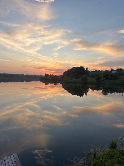 This is a vertical, eye-level, full-shot photograph of a tranquil lake at sunset, reflecting the colorful sky and surrounding landscape. The image depicts a peaceful natural scene, possibly in or near Kamysh, Russia, given the subtle hints of a rural or semi-rural environment.
The sky dominates the upper half of the frame, filled with wispy clouds painted in hues of pale orange, pink, and soft blue. These clouds are illuminated by the setting sun, which is partially visible behind a distant treeline. The treeline, a dark mass of foliage, stretches across the horizon, with hints of scattered buildings and utility poles suggesting a small settlement.
The lake's surface is remarkably still, creating a near-perfect mirror image of the sky and the distant shore. The reflections are clear, showing the vibrant colors of the sunset and the silhouetted trees. In the foreground, a wooden pier juts out from the lower left corner of the frame, with its weathered planks visible. To the right of the pier, the edge of the lake is lined with lush green grass and some aquatic vegetation, adding a touch of natural detail to the scene. The overall impression is one of serenity and natural beauty at the close of day. There are no people or animals visible in the image.