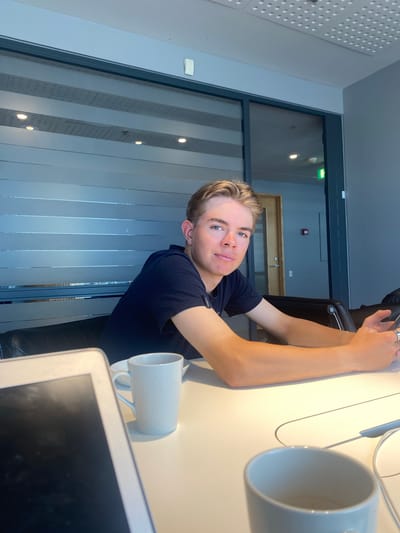 A young man with blond hair and blue eyes is seated at a desk, wearing a dark blue t-shirt. He is looking at the camera with a slight smile. On the desk, there are two white mugs and a laptop. The background features a window with vertical blinds and a door visible in the distance. The location is Kastrup, Denmark.