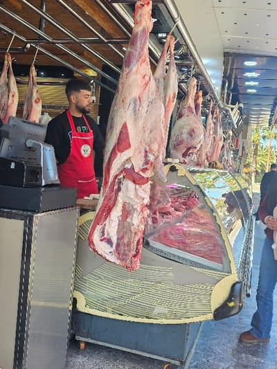 The scene depicts an open-front butcher shop in Boufakrane, Morocco. Multiple raw animal carcasses hang from metal hooks suspended from an overhead metal rail system. A large, skinned carcass dominates the central foreground, hanging above a curved glass refrigerated display case.

A male butcher, with dark hair and a beard, stands on the left behind a counter. He wears a black long-sleeved shirt under a red apron featuring a white logo. His gaze is directed towards the right side of the frame. On the counter in front of him are a black electronic scale and another black rectangular device.

The refrigerated display case beneath the hanging meat contains numerous cuts of raw red meat. The case's exterior is grey with a light yellow striped pattern.

To the far right, the lower legs and torso of a second individual are visible; this person wears blue jeans and brown shoes. The background, beyond the shop's structure, shows an outdoor environment with visible palm trees and ambient daylight, indicating a street-side or open-air market setting.