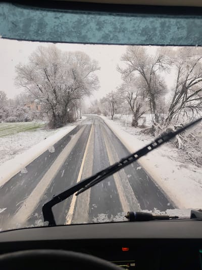 The image depicts a winter scene captured from inside a vehicle, looking out through a snow-covered windshield, in Klenovik, Bulgaria. Snowflakes are actively visible on the glass, and a black windshield wiper is positioned across the lower right portion of the view, indicating ongoing clearing. The dark dashboard is visible at the bottom, partially obscuring a steering wheel on the left. An object, appearing to be a tool or nozzle, rests horizontally on the dashboard to the right of the steering wheel. Dashboard indicators show an orange warning light and a digital display.

Outside the vehicle, a two-lane asphalt road stretches into the distance, partially cleared of snow, revealing sections of black asphalt and light-colored lane markings, which appear to be white and yellow. Snow blankets the shoulders of the road and the surrounding landscape. On both sides of the road, deciduous trees stand heavily coated in snow and frost. A green field is visible on the left side of the road, also partially covered in snow. A light-colored building is partially discernible behind the trees on the left side, further in the distance. The sky is overcast, indicating a snowy or heavily clouded day.