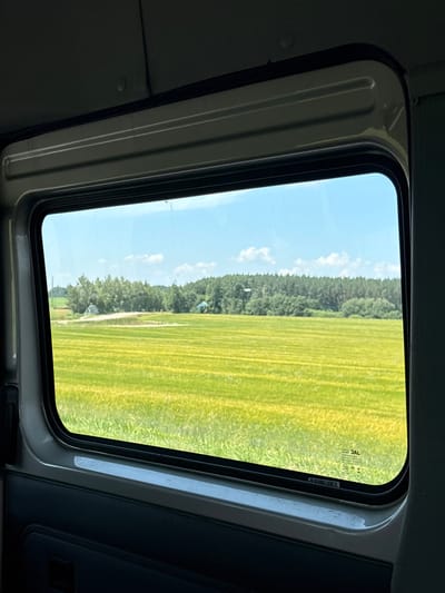 The image is an interior shot of a vehicle, looking upwards through a sunroof at a bright, sunny daytime scene. The sunroof frame, a dark grey color with some metallic trim, surrounds a view of a vibrant green field divided by lines, suggesting it might be a mowed lawn or a cultivated crop. A dense forest borders the field, with a clear blue sky dotted with a few white clouds above. The overall setting evokes a rural landscape, possibly in Belarus given the location context. There is no specific activity or event depicted, and no people are visible. Notable details include the clear weather and the overall brightness of the scene, indicating midday. The image also shows a small, white, possibly circular object suspended in the air over the field, its nature unclear. There is some faint text on the glass of the sunroof, appearing to be certifications or manufacturer marks, but it is largely illegible.