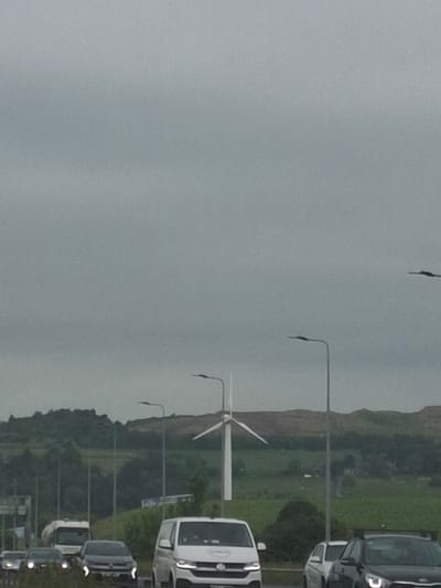 The image depicts a landscape scene with a wind turbine in the background, situated in a rural or semi-rural area that could be part of the city of Kirklees, United Kingdom. In the foreground, a road is visible with several vehicles, including a white van, a black car, and a truck. The sky is overcast, suggesting a cloudy day. The environment appears to be a typical highway setting with roadside lighting.