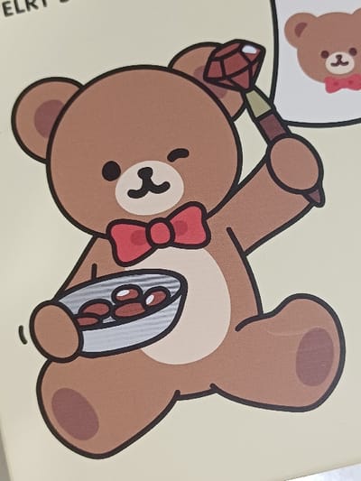 The image displays a graphic illustration printed on a light yellow surface, located in 서대문구, South Korea. The central subject is a cartoon teddy bear, rendered in brown with lighter tan for its snout and belly, and darker brown for paw pads and ear interiors. The bear possesses black dot eyes, a small black nose, and a subtle smile, with its right eye closed in a wink. A red bow tie is positioned at its neck. Its right arm is raised, holding an object with a brown handle and a prominent faceted red gem as its head. The left arm cradles a small, silver-colored bowl containing four distinct oval, reddish-brown objects. In the upper right quadrant of the image, a white thought bubble encloses a smaller, simplified illustration of a brown teddy bear, also wearing a red bow tie. Partial text reading "ELRY B" is visible in the upper left quadrant.