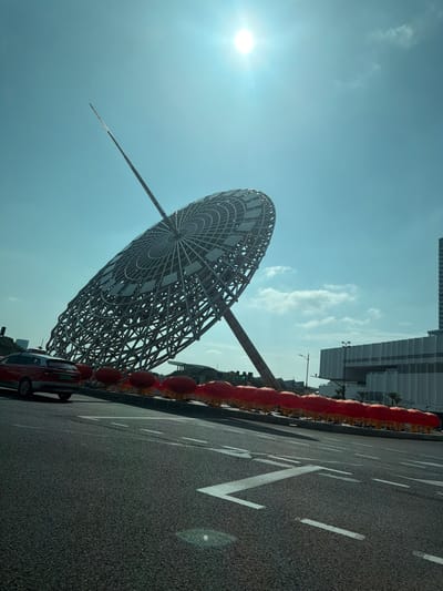 The image presents a sunlit urban scene in Yanqiao, China, under a bright blue sky with scattered clouds and a visible sun in the upper right, generating a lens flare. The primary subject is a large, contemporary, silver-colored lattice structure dominated by a tilted, disc-shaped array of square panels, supported by a central spire extending upwards.
In the foreground, an asphalt road displays white lane markings, including solid lines, dashed lines, and a distinct 'Z' shape, angled slightly from the lower left to the right. A red SUV or crossover vehicle, possibly a Haval H6, is partially visible on the left side of the road, oriented away from the camera.
Adjacent to the road and beneath the large structure, a uniform row of approximately 20-25 red, spherical lanterns is arranged along a curb or median. In the background, modern multi-story buildings, characterized by white and grey facades, are visible, along with distant green traffic lights on poles. No individuals are present in the frame.