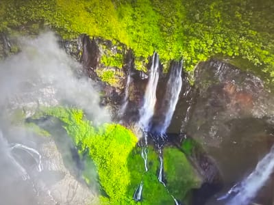 The image shows a view of a waterfall from above. The waterfall cascades over a cliff face that is covered in lush green vegetation. Mist is present around the waterfall. There is no indication of Basingstoke and Deane in the image. There are no people, subjects, objects, actions, or interactions to be described other than the waterfall and surrounding environment.