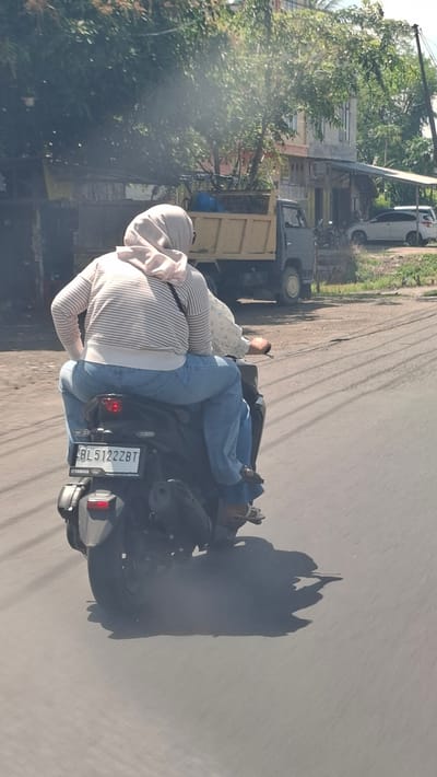 Two people are riding on a motorcycle on a paved road in what appears to be a rural or suburban area in Cotbadatunong, Indonesia. The rider is wearing blue jeans and a blue shirt. The passenger is wearing a light-colored headscarf and a striped shirt. Both are wearing casual clothing.
In the background, there is a light-colored truck with a yellow bed, and several buildings. Trees and foliage are visible on the left side of the image. The road is paved and has tire tracks, indicating it is a frequently used thoroughfare. The lighting suggests it is daytime, possibly late morning or early afternoon, with sunlight casting shadows on the road. The weather appears to be clear and sunny.
A license plate on the back of the motorcycle is visible and reads "BL 5122 ZBT". A Yamaha logo is also visible on the back of the motorcycle. The overall scene depicts everyday transportation and life in a populated area.