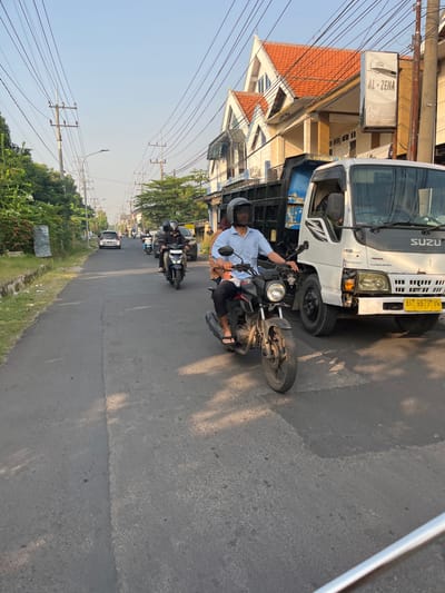 This is a street scene in Sugih Waras, Indonesia, featuring a busy road with several vehicles. In the foreground, a man wearing a light blue shirt and a black helmet is riding a black motorcycle. He is on the right side of the road, with a large white truck partially obscuring him from view. The truck has "SUZUKI" written on its side. Behind the man on the motorcycle, other motorcycles and a silver car are visible further down the road. The street is lined with trees and utility poles, and buildings are present on the right side of the image. The sky is clear and blue, indicating daylight. The overall scene depicts typical traffic conditions in a developing urban or suburban area.