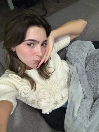 A woman reclines on a gray couch, draped in a fluffy gray blanket. She wears an off-white sweater adorned with raised floral designs. Her dark brown hair frames her face as she gazes upward. Her left hand is positioned near her cheek, fingers slightly touching her face. Her skin is fair with a hint of blush. The overall composition suggests a relaxed, intimate moment.