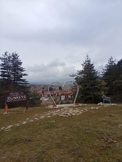 This image depicts an elevated outdoor scene overlooking the village of Madzhare, Bulgaria, within the area of Rila National Park, under an overcast sky.

The foreground consists of a grassy slope with numerous patches of exposed soil and several clusters of dark animal droppings. An irregular path of light-colored flat stones is partially visible, leading towards the center of the frame.

In the midground, from left to right, are three main subjects:
1.  A wooden signpost, dark brown with white lettering, states "WELCOME to Happy DONKEYS" above "Madzhare, Rila National Park." A small orange carrot cutout is affixed to the lower left of the sign.
2.  A large, white, heart-shaped metal structure, potentially an illuminated display, is positioned centrally on a low concrete base with red accents.
3.  A life-sized, grey-blue statue of a donkey stands on a small green and white base to the right.
Several tall, dark green coniferous trees are situated behind and around these features, partially obscuring the view of the background.

The background features the village of Madzhare, comprising multiple buildings with distinctive red-tiled roofs and light-colored walls, nestled in a lower elevation. Beyond the village, tree-covered hills or mountains extend into the distance. The sky is uniformly grey and overcast. A small, dark, unidentifiable object is present in the upper left quadrant of the sky. No individuals are visible in the scene.