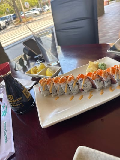 A high-angle interior photograph depicts a sushi meal served on a dark brown wooden table within a restaurant in Naples, United States. The central focus is a long, off-white rectangular plate holding ten pieces of sushi. Each sushi piece contains white rice, a dark red interior (consistent with tuna), is topped with orange roe, and is drizzled with a light orange sauce. The plate also features a small mound of pickled ginger and a dollop of green wasabi. To the left, a dark brown Kikkoman soy sauce bottle with a red cap stands beside a white paper sleeve labeled "Bamboo Chopsticks." Further left, a white dish with two compartments holds lemon wedges. Behind these items, a clear glass of water containing a white straw, a dark-colored smartphone, and dark sunglasses are present on the table. In the blurred background, a dark gray chair back is visible, with a large window revealing a sunlit exterior street scene featuring a sidewalk, parked vehicles, a road, and trees. No individuals are present in the frame.