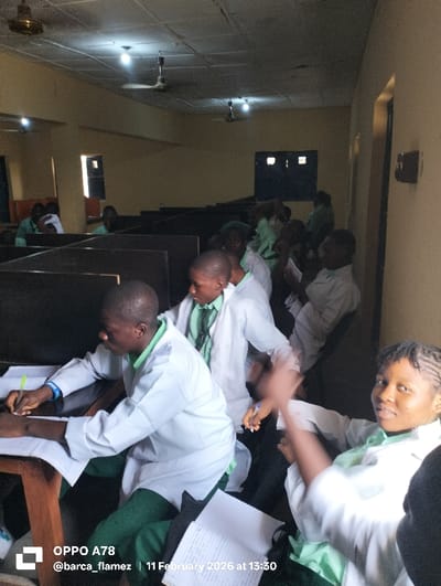 The image depicts an interior scene within a facility identified as being in Rinze, Nigeria, functioning as a classroom or study hall. The room features light-colored walls and a tiled ceiling from which multiple bare light bulbs and ceiling fans are suspended for illumination and air circulation.
Approximately fifteen to twenty individuals, primarily young adults, are present, all uniformly dressed in green collared shirts worn under white lab coats. In the immediate foreground on the left, a male is seated at a wooden desk, actively writing in an open notebook with a pen. A blue bracelet is visible on his left wrist. To his right, a female with dark braided hair is partially visible, looking directly towards the camera with a slight smile, her hands appearing blurred in motion as she holds a pen, with papers on the desk in front of her.
In the mid-ground and background, other individuals are seated at similar wooden desks, some of which are separated by dark partitions, suggesting individual workstations. The overall activity indicates a learning or study session. A window providing external daylight is visible on the left wall of the room, and a dark-framed window is present in the central background.