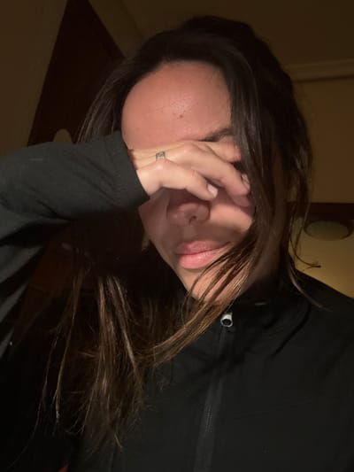 A woman with long, dark brown hair is seen from a close, angled perspective. Her face is partially obscured by her hand, which bears a ring. She wears a black long-sleeved shirt or jacket with a visible zipper. The lighting is dim, casting shadows across her face and clothing. A portion of a door is visible in the background, adding depth to the image.