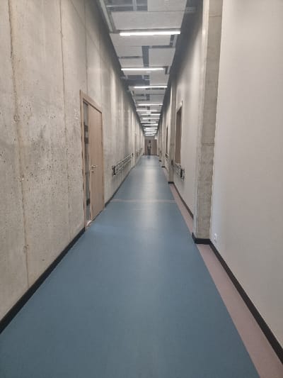 The image displays a long, well-lit, modern indoor corridor in Jaunmārupe, Latvia. The left wall is constructed from raw, textured concrete, featuring a light wooden door marked "E21" in the foreground and a metal handrail extending along its length. The right wall is smooth and light-colored, with a concrete support column prominent in the foreground and a series of three similar light wooden doors, each with a corresponding metal handrail, visible further down the hallway. The floor is a consistent blue-grey material, bordered by a black baseboard on the left and a narrow beige-pink strip above a black baseboard on the right. The ceiling consists of a suspended metal grid system holding light grey acoustic panels and numerous rectangular LED strip lights providing illumination. A single, indistinct individual, dressed in a dark top and light-colored bottoms, is visible standing motionless at the far end of the corridor, facing away from the camera.